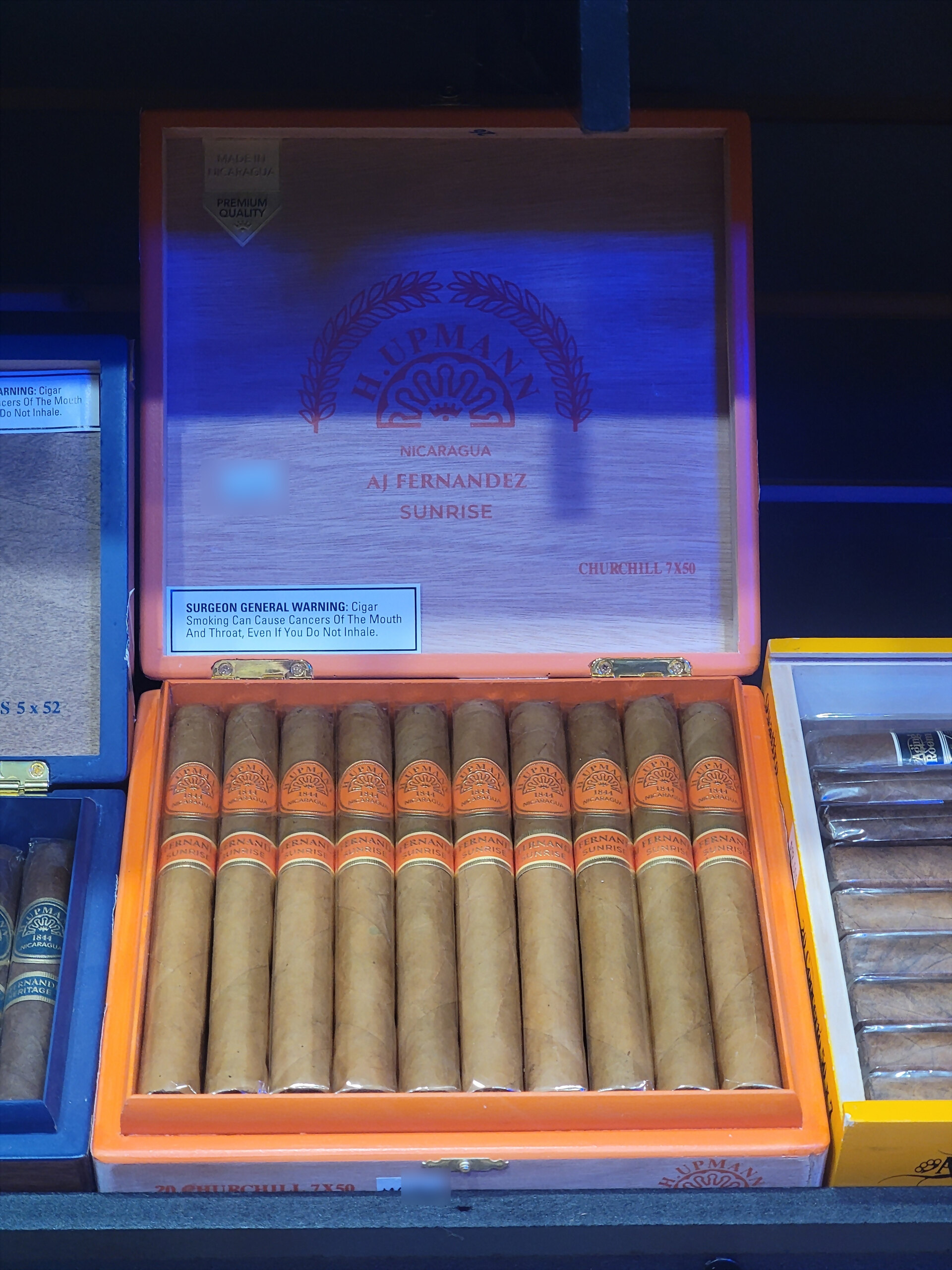 An H. Upmann Nicaragua by AJ Fernandez Sunrise box open on the shelf, CHURCHILL 7X50 on the inner lid