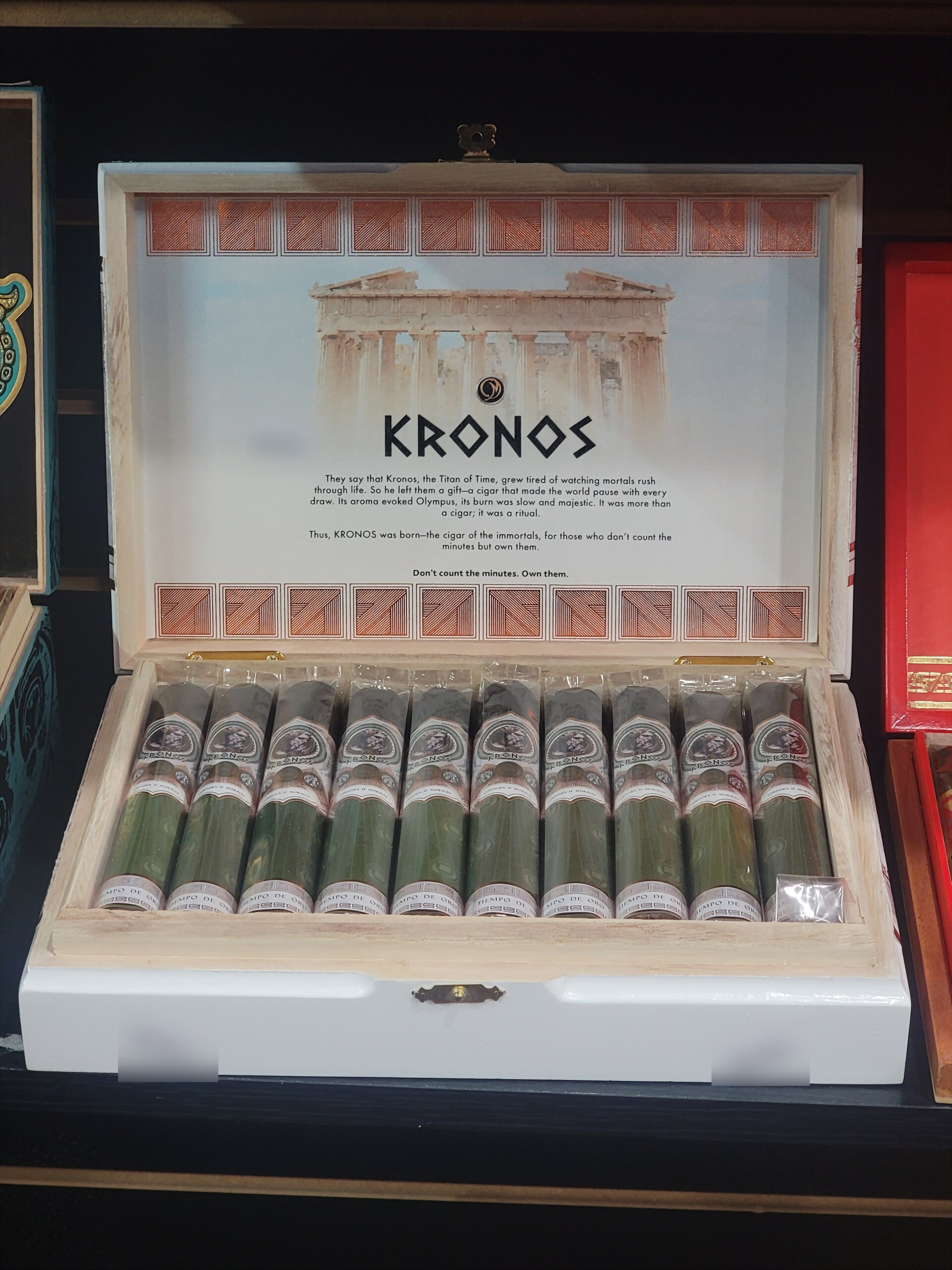 A Kronos box open on the shelf, the Parthenon printed across the inner lid above green-footed cigars